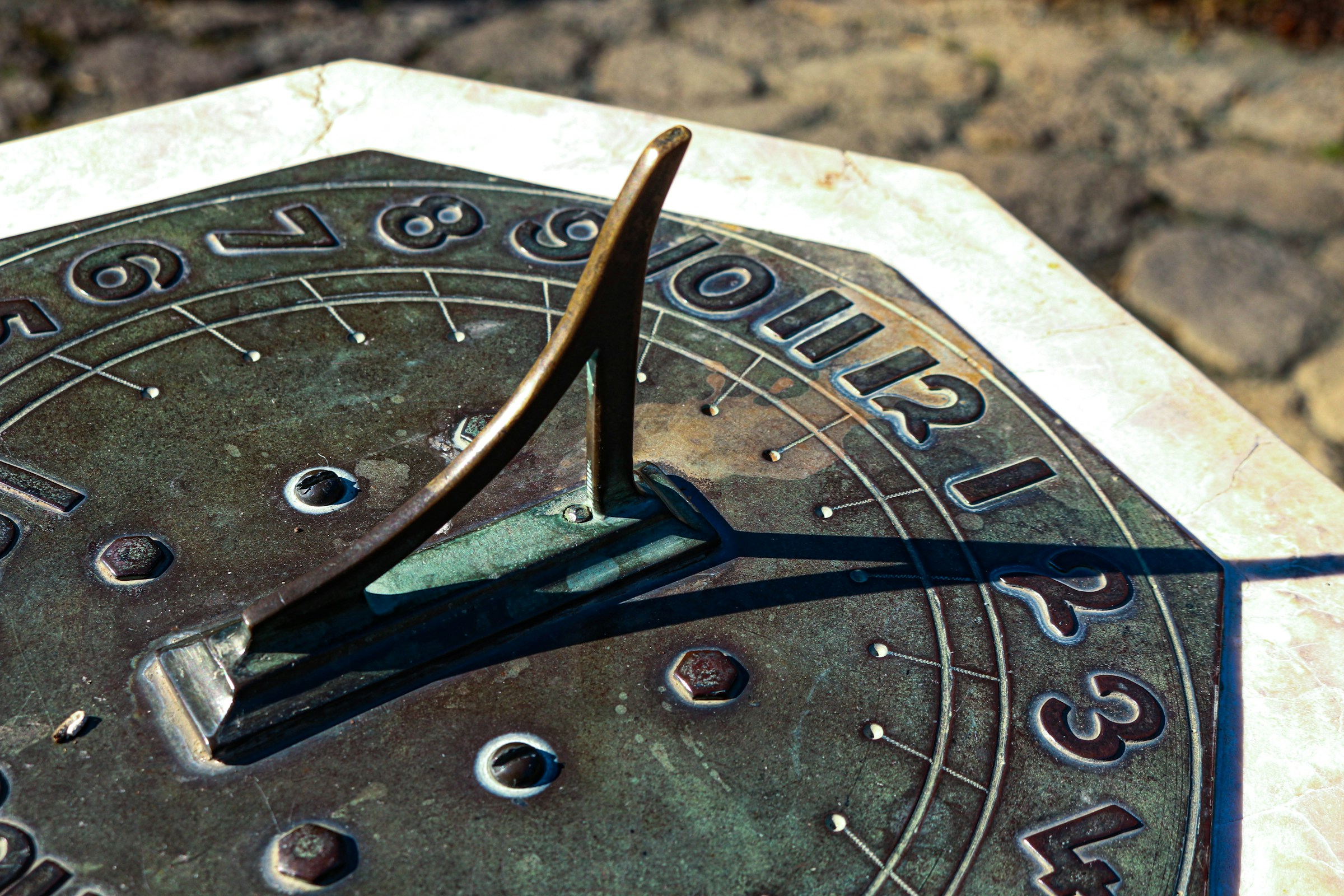 A close-up photograph of a bronze or copper sundial mounted on stone with the shadow pointing to two o'clock.